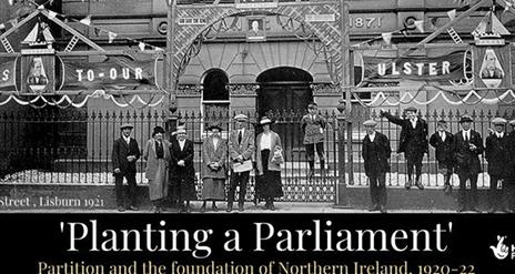 Image shows a black and white photo of group of men, women and children from the 1920s standing outside the gates of a building