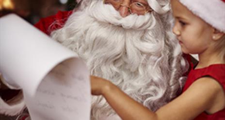 image shows picture of santa claus and a little girl in a red and white santa hat and red dress, looking at santas long list on a scroll of white pape