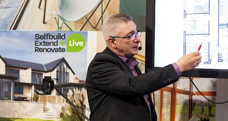 Image is of exhibitor pointing at a set of house plans at the Selfbuild Extend Renovate exhibition in Eikon Exhibition Centre Lisburn