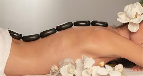 Image is of a lady with stones on her back for a hot stone massage. A display of white orchids are also in the image