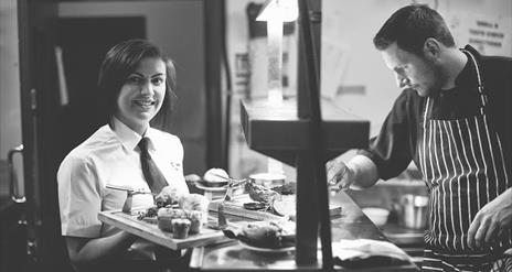 A black and white image showing the chef in the kitchen with a waitress carrying food.