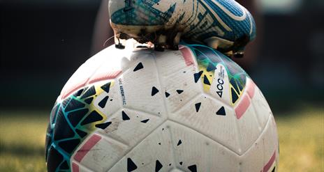 An image of a football with someone holding it in place by their football boot
