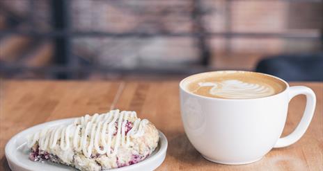 Image shows a white cup of latte and white side plate with a  traybake covered in icing.