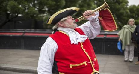 Image shows Town Crier dressed in traditional dress with red waistcoat and black hat.