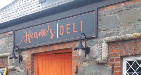 Image shows picture of the front of Keanes Deli stone building and part of the orange front door.  The signage has a grey background with orange colou
