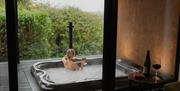 Person relaxing in an outdoor hot tub holding a drink beside a window view of greenery.