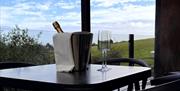 Table with ice bucket and champagne flutes on a glass enclosed balcony overlooking green hills.