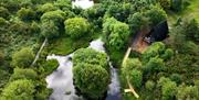 Aerial view of a luxury A-frame lodge nestled among woodland, with ponds, walking paths and green countryside surrounding the retreat.