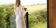 Person in a robe holding a drink on the balcony overlooking fields.