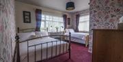 Image shows a double and single bed with two large windows, side table and lamps, chest of drawers in foreground