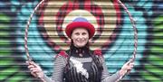 Woman in costume holding a hula hoop in front of a painted door. Circusful performer. Friday Fun in the Park. Moira Demesne.