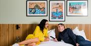 Image shows a man and woman lounging on a double bed with paintings of Northern Ireland on the background wall