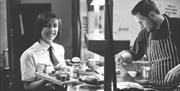 A black and white image showing the chef in the kitchen with a waitress carrying food.
