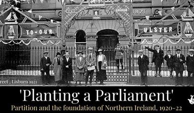 Image shows a black and white photo of group of men, women and children from the 1920s standing outside the gates of a building