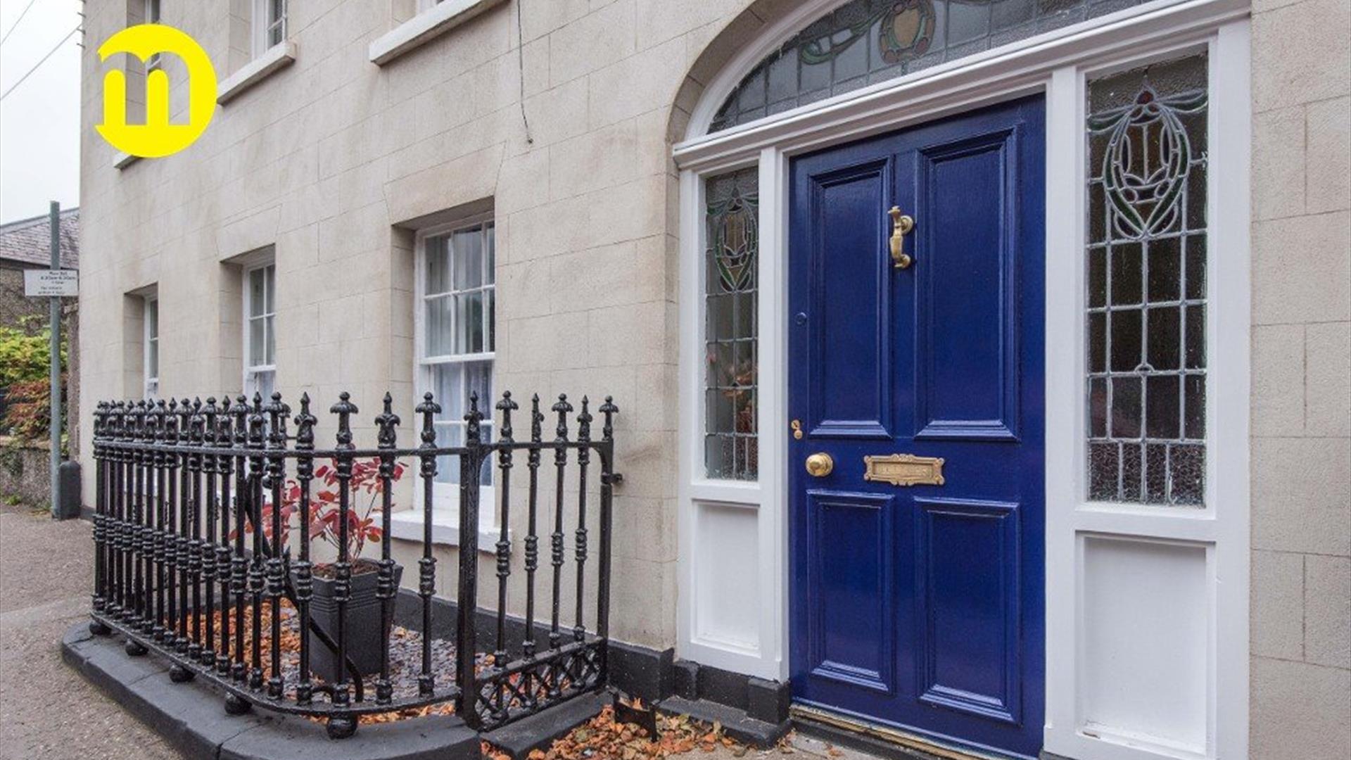 Image shows royal blue front door with iron railing to the left of the door Door leads out onto footpath on street