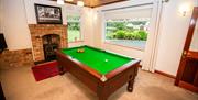 Image shows room with a pool table, TV on wall, wood burner in fireplace and large window looking out onto driveway
