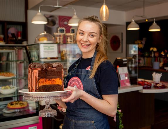 Image shows smiling member of staff holding a large  chocolate and orange cake