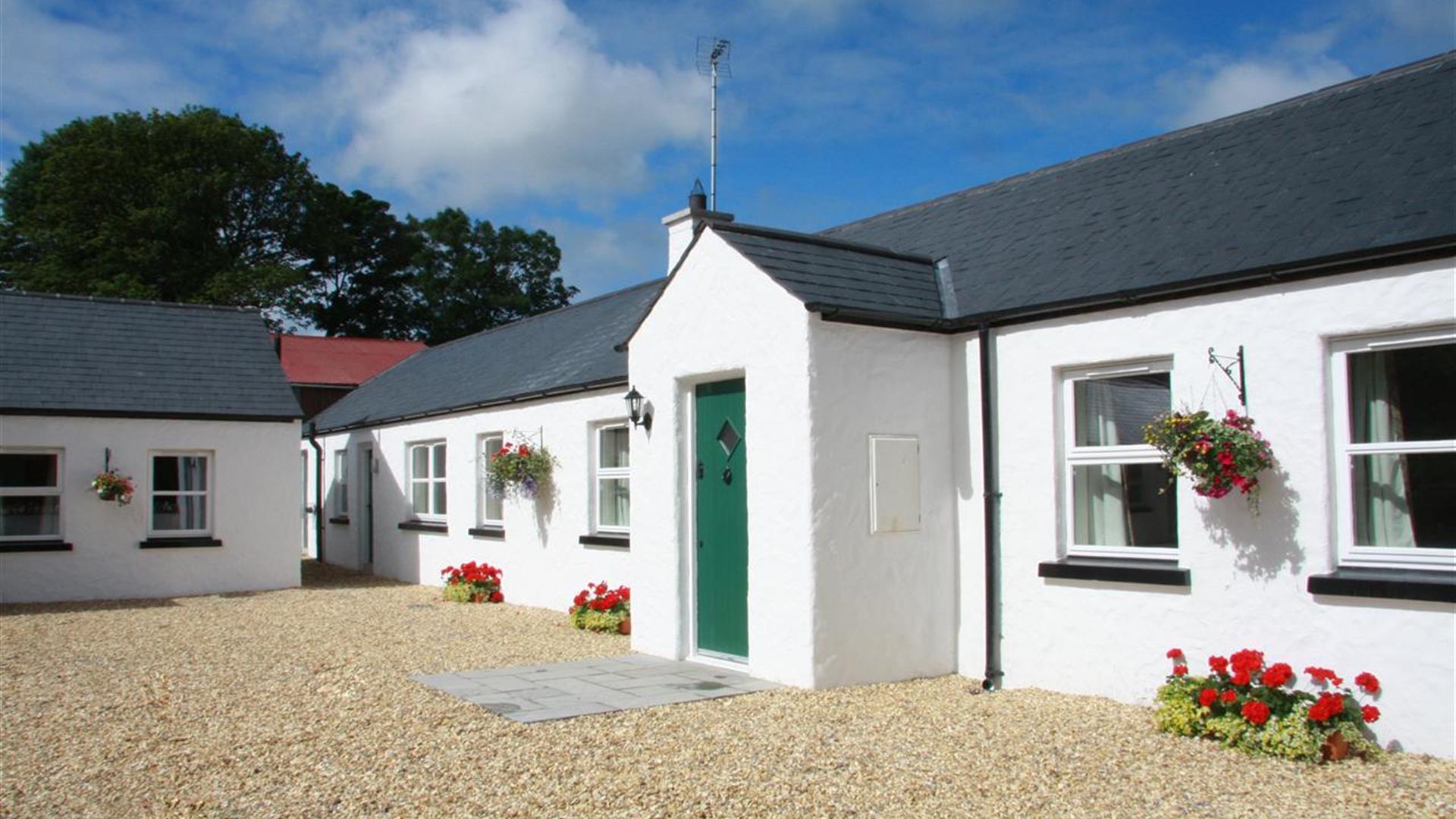 Image shows front of whitewashed property with green door and gravel drive. Flowerbeds on gravel outside windows