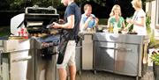 Image shows a man barbecuing with people enjoying food in the garden