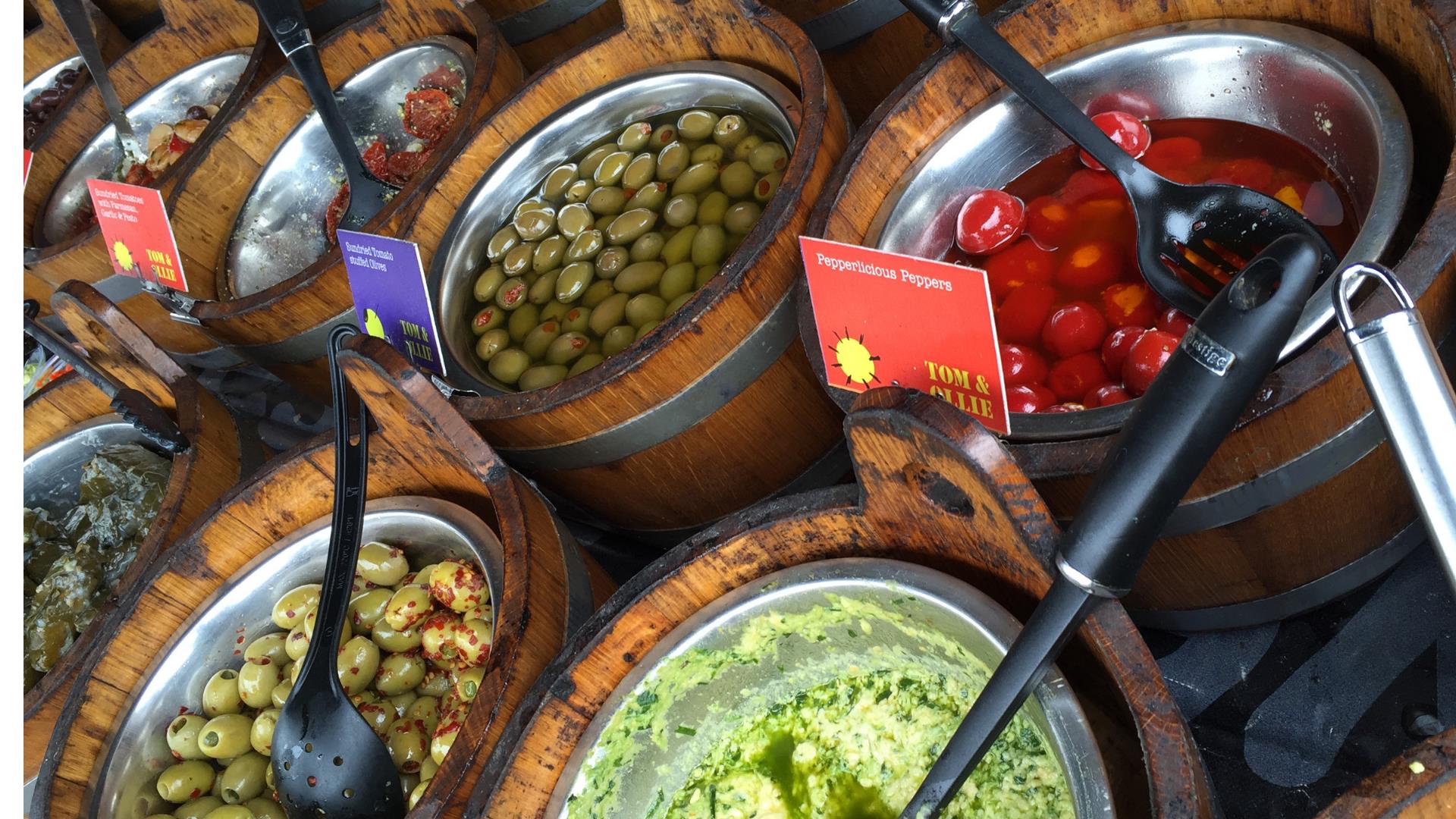 Image shows wooden tubs of various food stuffs such as olives and cherries