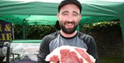 Image shows a smiling man holding up a large cut of beef at his stall