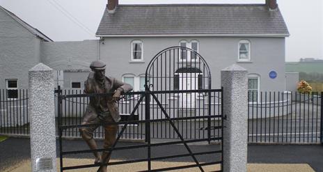 Image is of Harry Ferguson bronze statue leaning on gate  in front of his family home