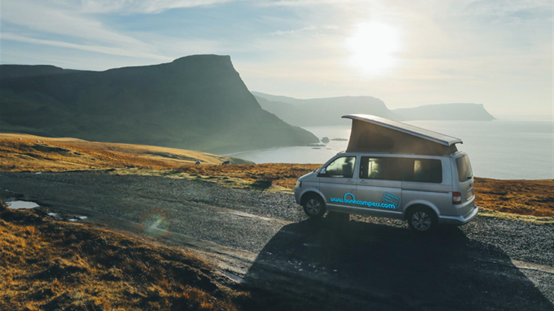 Image is of campervan driving down a road with sea and mountains in view