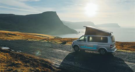 Image is of campervan driving down a road with sea and mountains in view