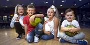 Image is of children sitting on the ground holding bowling bowls inside the bowling alley