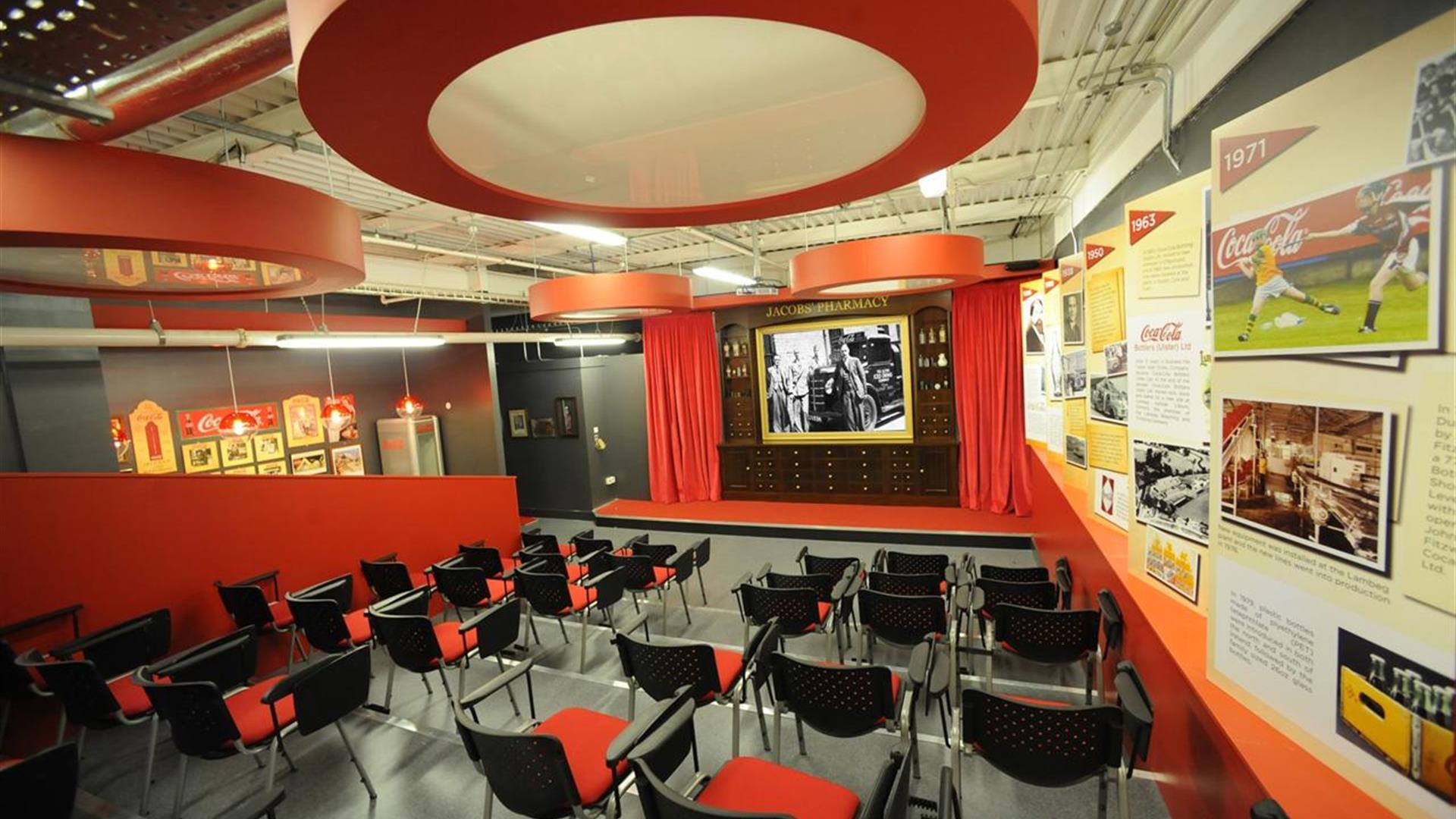 Image is of a meeting room with lots of chairs, images on the walls and large film screen on a stage