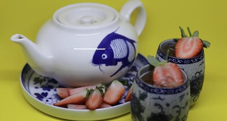 Image is of a teapot with a fish on it and some strawberries in a small vase