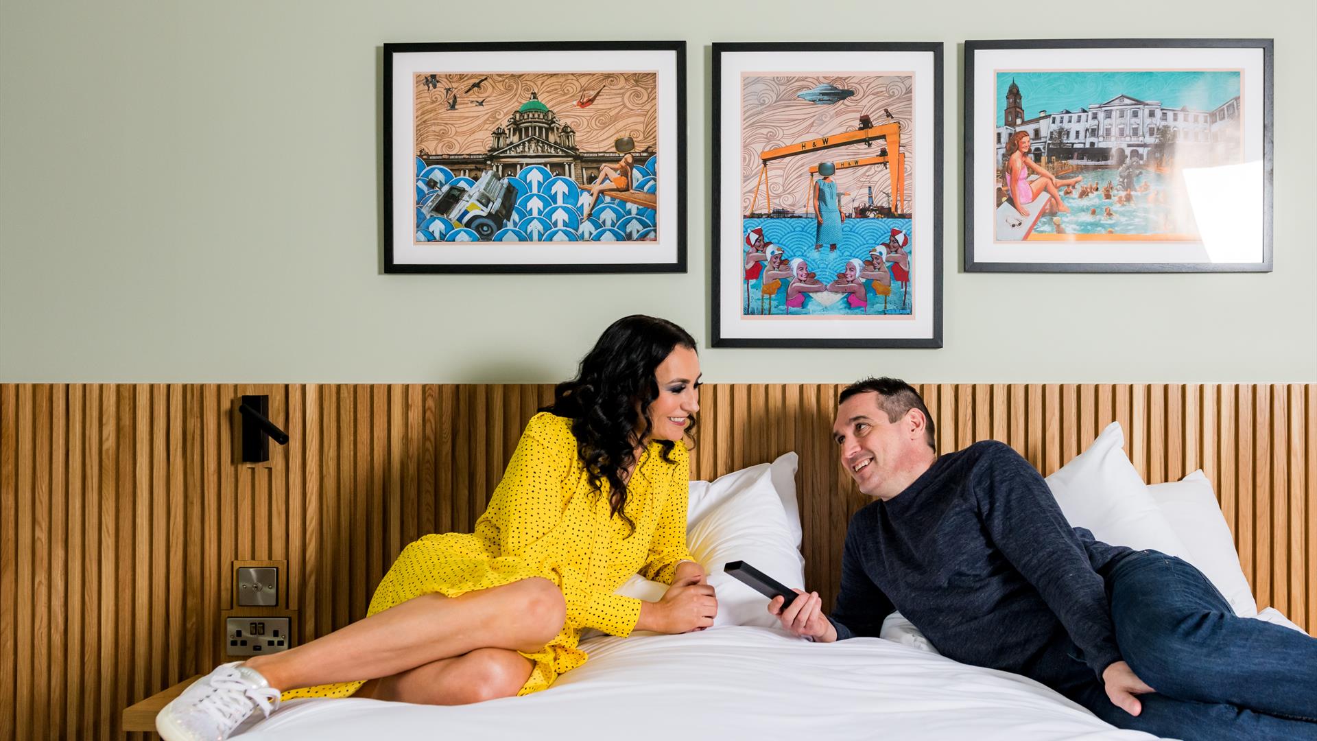 Image shows a man and woman lounging on a double bed with paintings of Northern Ireland on the background wall