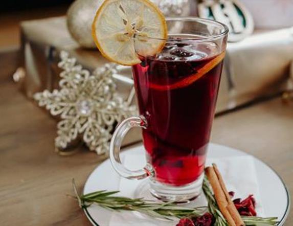 Image shows glass mug of mulled wine with dressing and cinnamon sticks and garnish on the side plate with silver decorations in the background of the