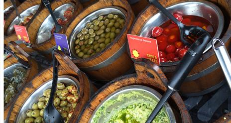 Image shows wooden barrels of different foods such as olives at the market