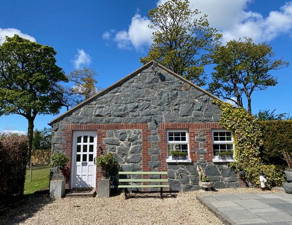 Image shows stone cottage covered in ivy at the right hand side plus white front door and bench outside