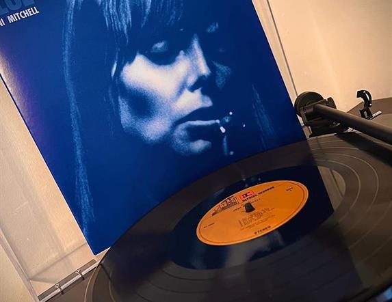 Picture of Joni Mitchell on the cover of her album in the background with a vinyl record on record player to the fore