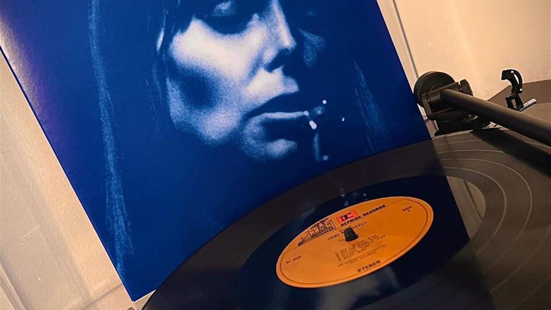 Picture of Joni Mitchell on the cover of her album in the background with a vinyl record on record player to the fore