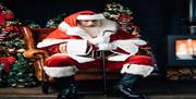 Image is of Father Christmas sitting beside Christmas tree and warm stove