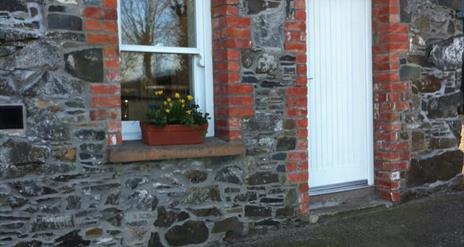 Image is of a stone cottage with a white front door and flowerbed on the window sill