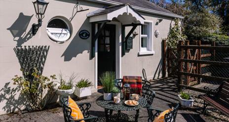 Image is of patio area at back of cottage with wrought iron table and 2 chairs.