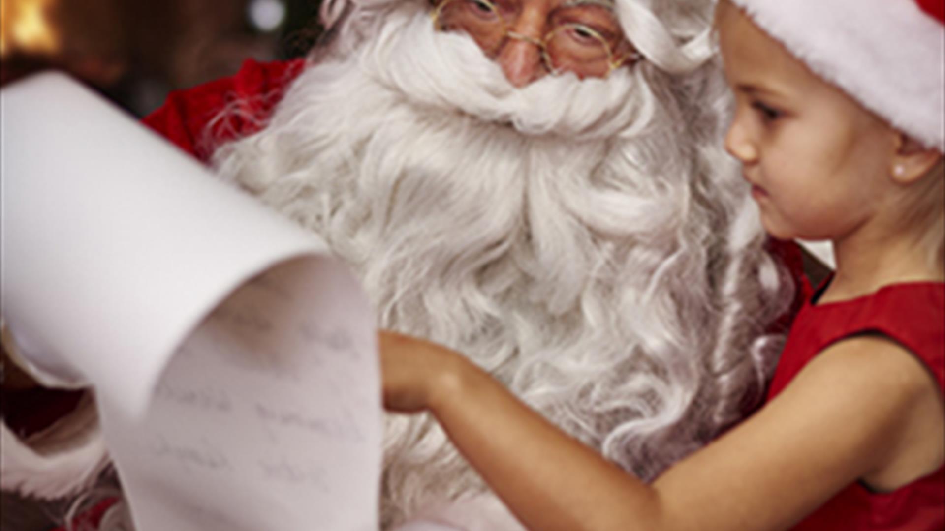 image shows picture of santa claus and a little girl in a red and white santa hat and red dress, looking at santas long list on a scroll of white pape
