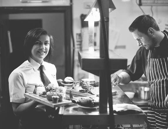 A black and white image showing the chef in the kitchen with a waitress carrying food.