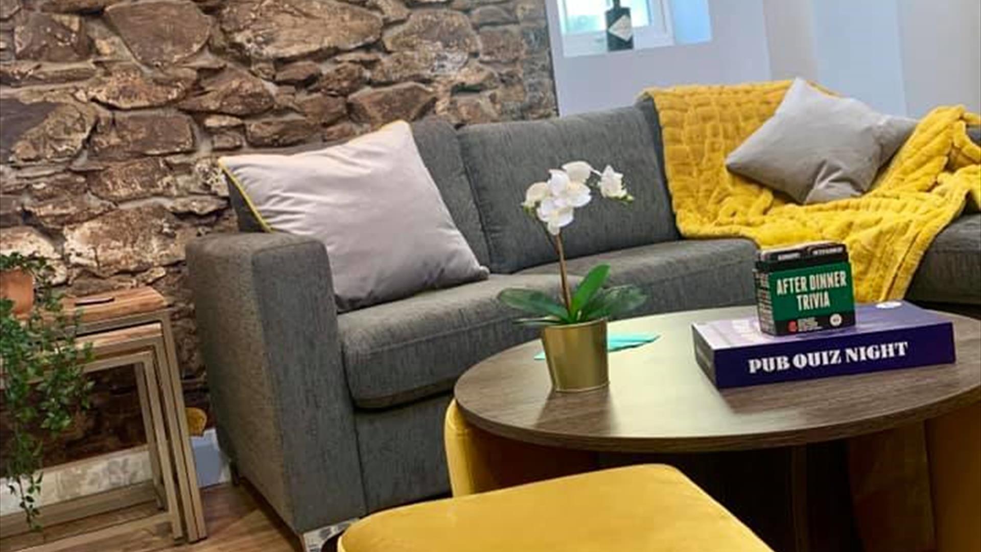 Image shows lounge area with sofa, foot stool, exposed brick wall with 2 clocks showing the time in Comber and New York. Wooden flooring and coffee ta