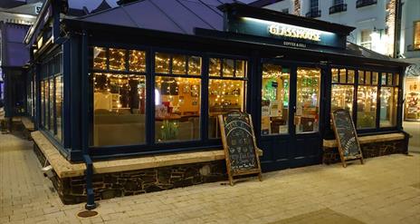 Image shows picture of the front of the glasshouse cafe 
windows with lights inside and two chalkboards leaning against premises.