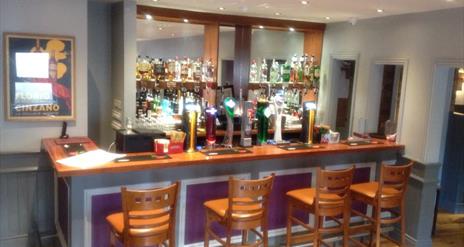 Image shows bar area with 4 bar stools on wooden bar bench and bar is painted a soft lilac colour with recessed lighting overhead.
