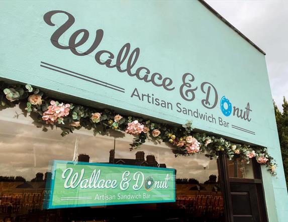Image shows a pale blue front sign of Wallace & Donut cafe premises in Lisburn with a pink flower display across the top of the window.